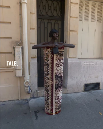 An African American female fashion model presents an abstract carpet-dress in a street setting on a sunny day, complemented by a small, black handbag.