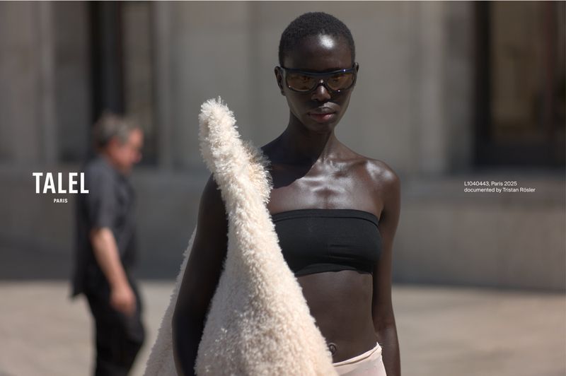 Fashion shot of an African-American woman in sunglasses and tube top, carrying a fuzzy bag outdoors with a man visible in the background.