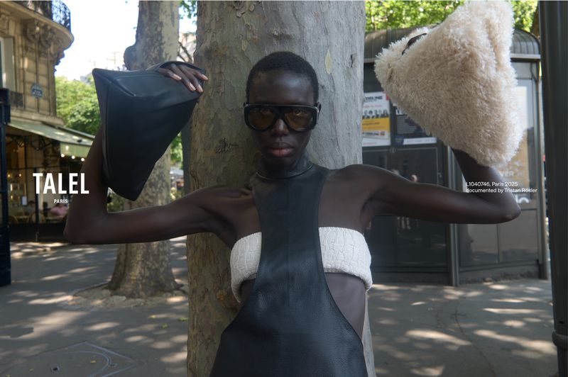 Fashion shot of a Black woman wearing a black dress, sunglasses, and holding two purses in an outdoor urban setting with greenery.