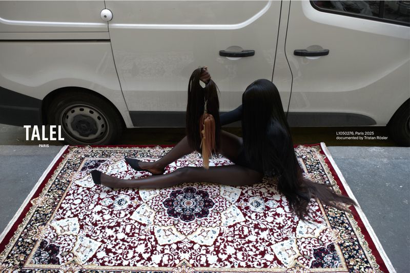 Woman in full body suit poses on patterned rug with hair extensions and van in the background, creating a surreal fashion image.