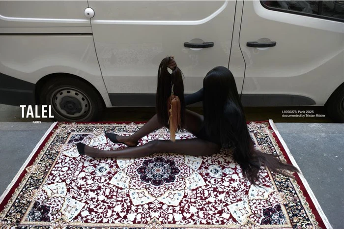 Model seated on an ornate patterned rug outdoors holding hair extensions and a handbag next to a white van