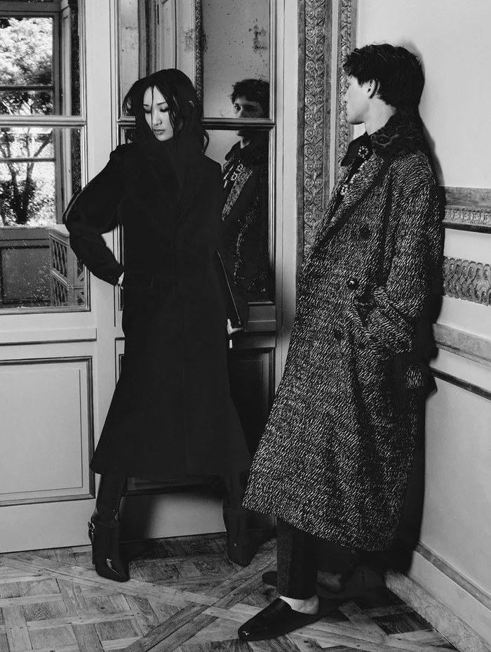 Black and white editorial portrait of two models in structured coats posing inside an ornate room