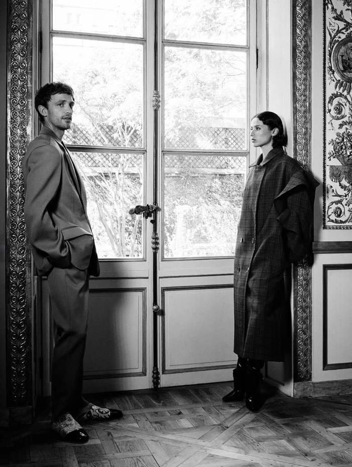 Monochrome fashion editorial image of a man in a suit and a woman in a long plaid coat standing in an ornate room
