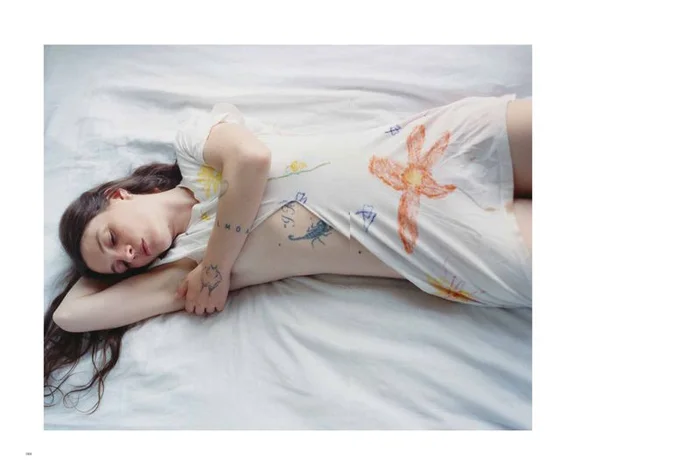 A woman with long dark hair lying on a white bed wearing a white dress with colorful crayon illustrations
