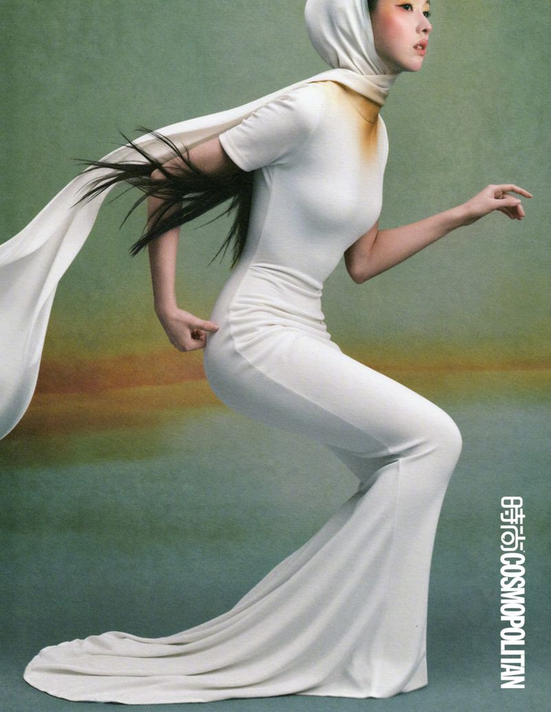 Woman in a white hooded dress with a long trailing scarf posing against a textured green background in a fashion editorial.