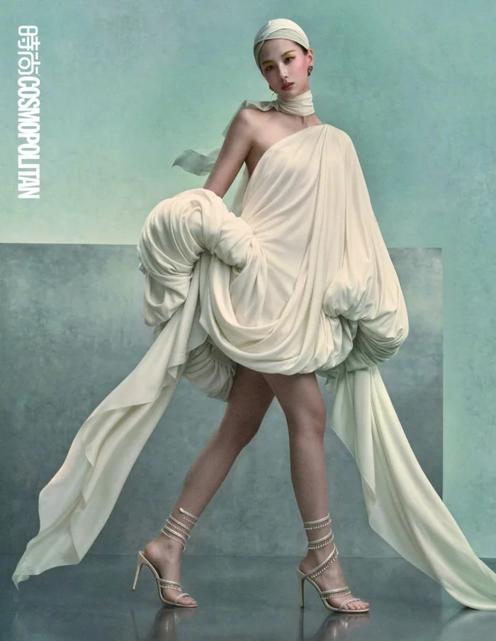 Woman in draping cream dress and matching headwrap with high heels standing against a textured green studio backdrop.