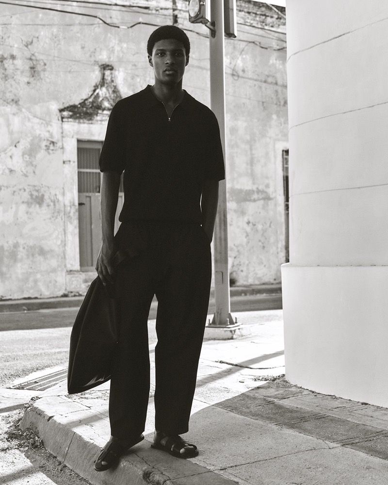 Monochrome outdoor portrait of a Black adult male model in a polo shirt and trousers, holding a jacket, standing on a city street.