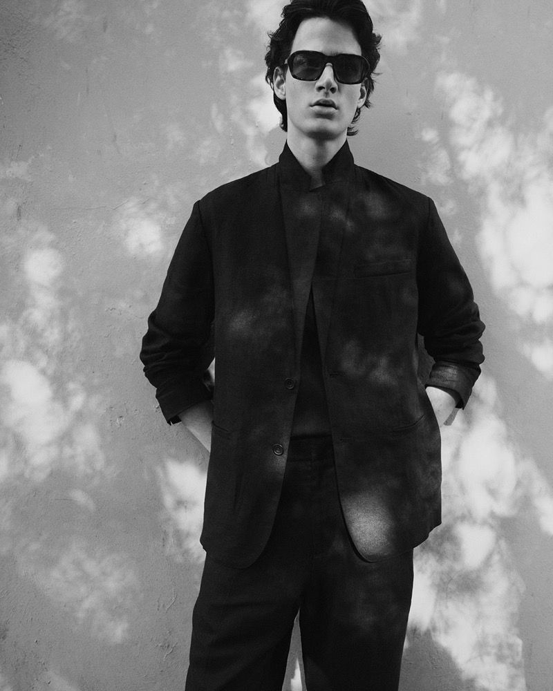 A fashionable male model stands in a blazer and trousers, wearing sunglasses, posing against a wall with leaf shadows.