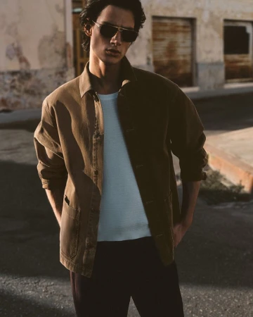 A young adult male fashion model poses outdoors wearing aviator sunglasses, an olive-green jacket, a t-shirt, and dark pants.