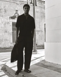 Black and white portrait of a black man wearing a dark polo shirt and trousers standing on an urban street.
