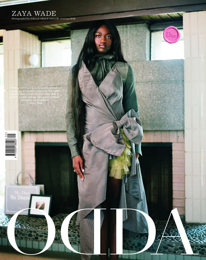 Zaya Wade standing indoors wearing a grey asymmetric dress with a large bow over a green ruffled top for ODDA Magazine.