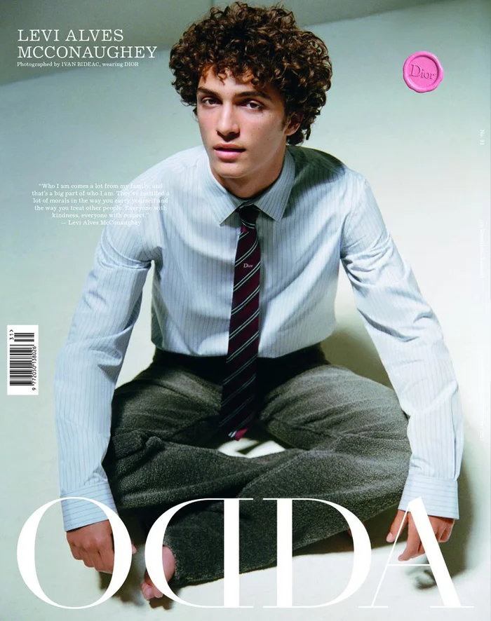 Levi Alves McConaughey seated cross-legged on a white studio floor wearing a light blue pinstripe shirt, dark tie, and grey trousers.