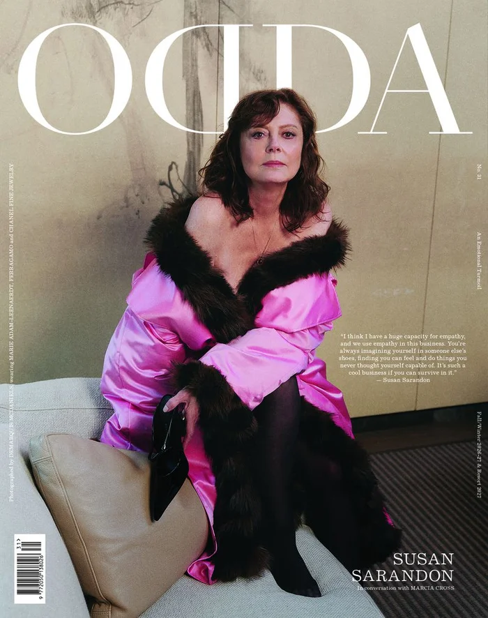 Susan Sarandon seated on a sofa wearing a bright pink satin coat with dark fur trim for ODDA Magazine