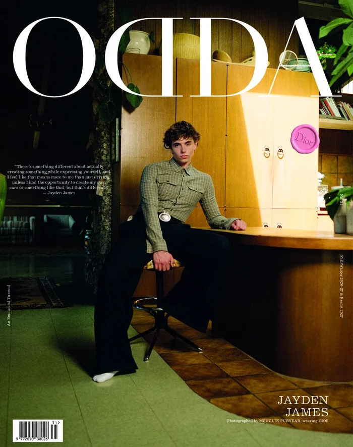 Young man in a patterned green shirt seated on a stool inside a domestic interior with wooden cabinetry and green flooring.