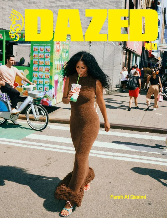 Woman with long dark curly hair wearing a brown knitted dress drinking from a cup while walking on a city street.