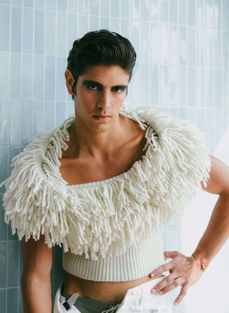 Headshot of a light-skinned male model with slicked-back dark hair wearing a fringed sweater and light makeup against a blue tile wall.