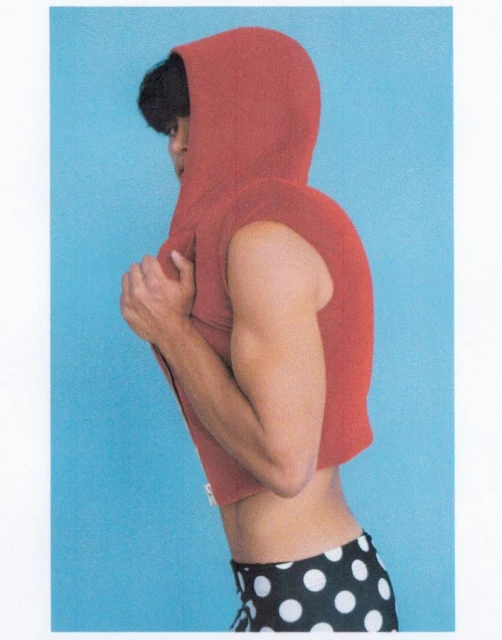 Side profile of a muscular adult with dark hair, wearing a red sleeveless hoodie and black and white polka dot shorts, against a blue background.