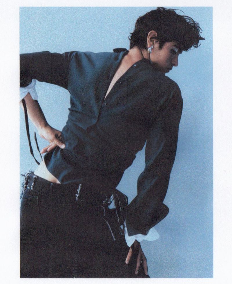 A person in a black button-down shirt and ripped jeans poses with their back arched against a pale blue background in a fashion shot.