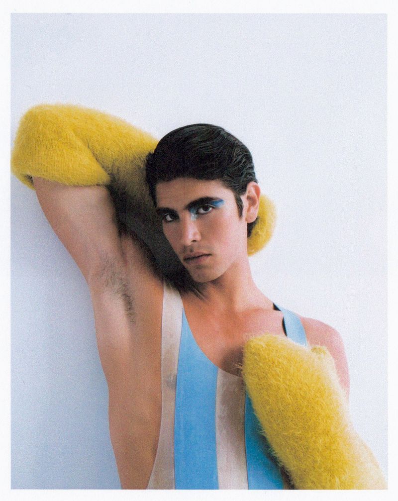 Fashion portrait of a young male model with dark hair and dramatic makeup, wearing a striped top and yellow furry accessories against a white background.