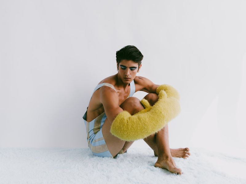 Young adult male with makeup in studio, wearing striped outfit and yellow fuzzy sleeves sitting on white rug against white background.