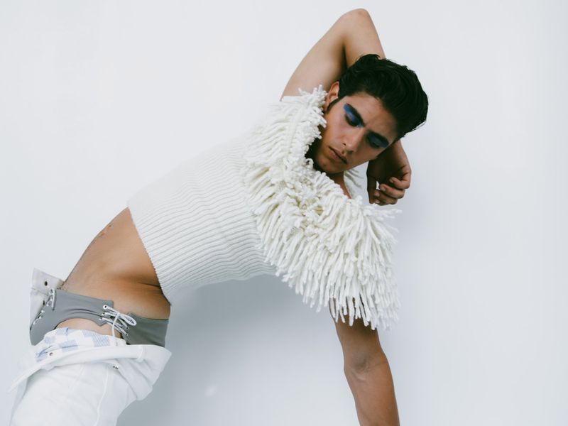 Person in white sweater poses against white background. Fashion shot featuring sweater with fringe detail and blue eyeshadow.