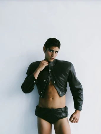 Man poses in black leather jacket and briefs against a white background, fashion forward with striking visual.