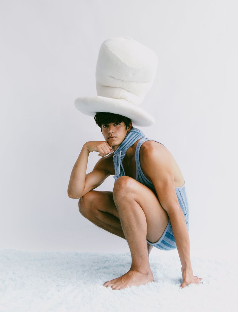 Asian male model squats wearing a blue striped outfit and oversized white hat against a plain white backdrop, striking a playful pose.