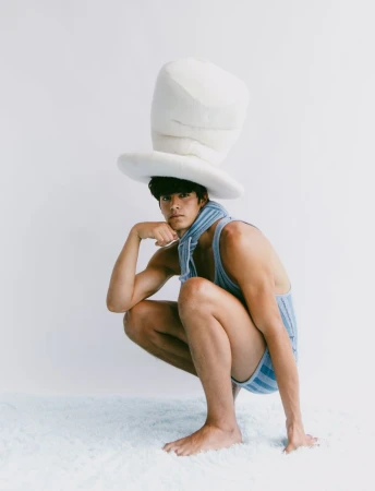 Asian male model squats wearing a blue striped outfit and oversized white hat against a plain white backdrop, striking a playful pose.