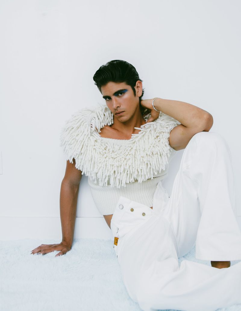 Stylish male fashion model poses in white outfit with textural fringe shoulder piece, sitting against a clean white studio background.