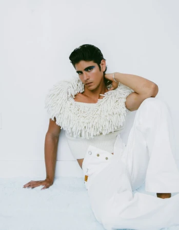 Stylish male fashion model poses in white outfit with textural fringe shoulder piece, sitting against a clean white studio background.