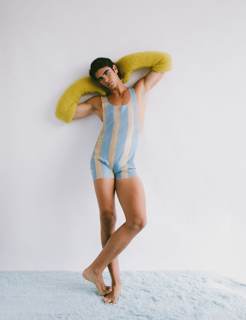 Full-length studio shot of a bare-chested adult male in a vintage striped swimsuit with fuzzy yellow shoulder pads, leaning against a white wall.