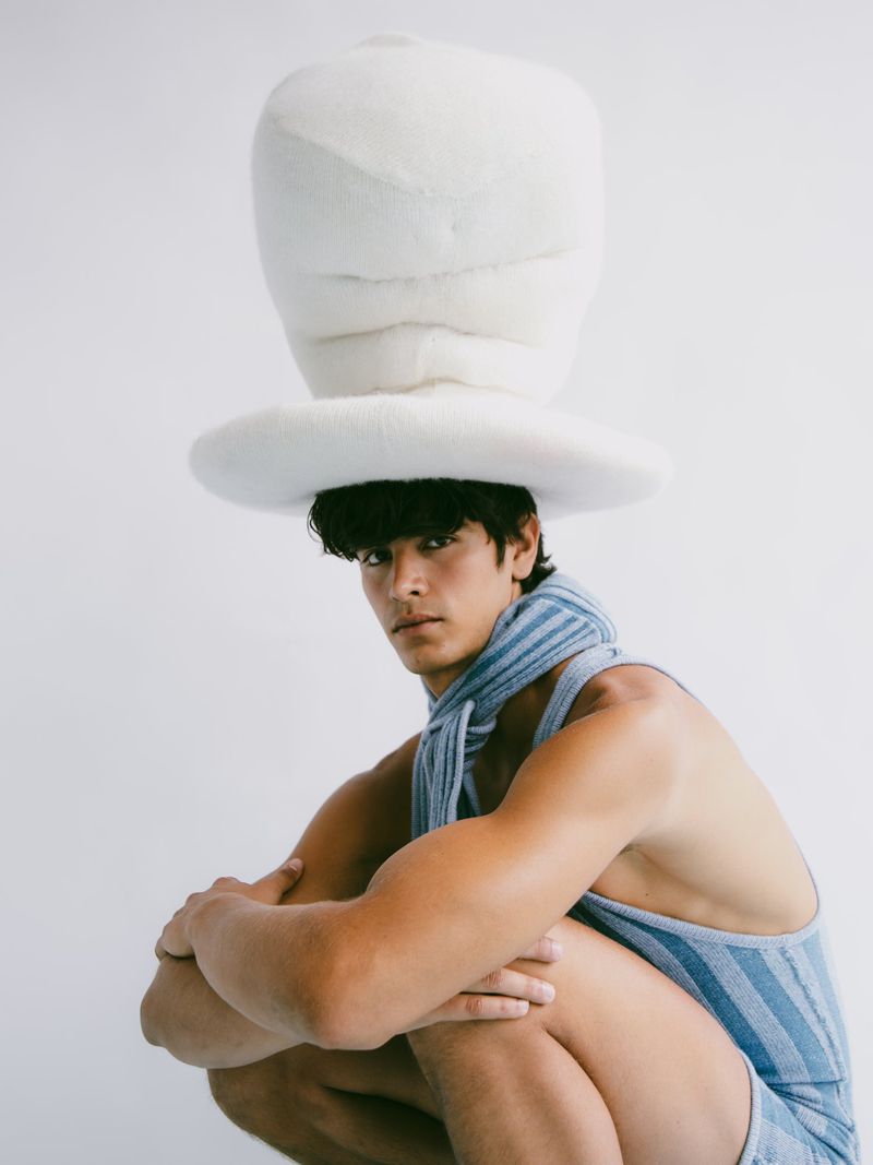 Man wearing a large, white fuzzy hat and blue striped outfit poses in front of a white background, looking at the camera.