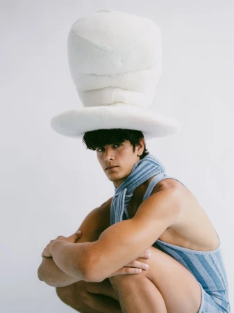 Man wearing a large, white fuzzy hat and blue striped outfit poses in front of a white background, looking at the camera.