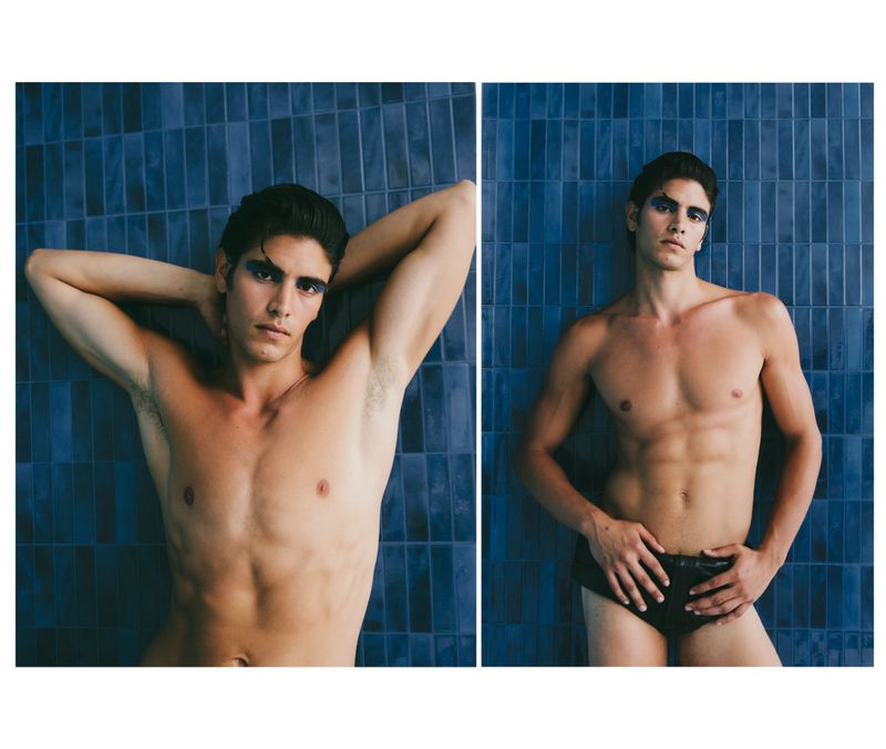 A male fashion model poses shirtless in front of a blue-tiled wall in a studio setting, showcasing eye makeup.