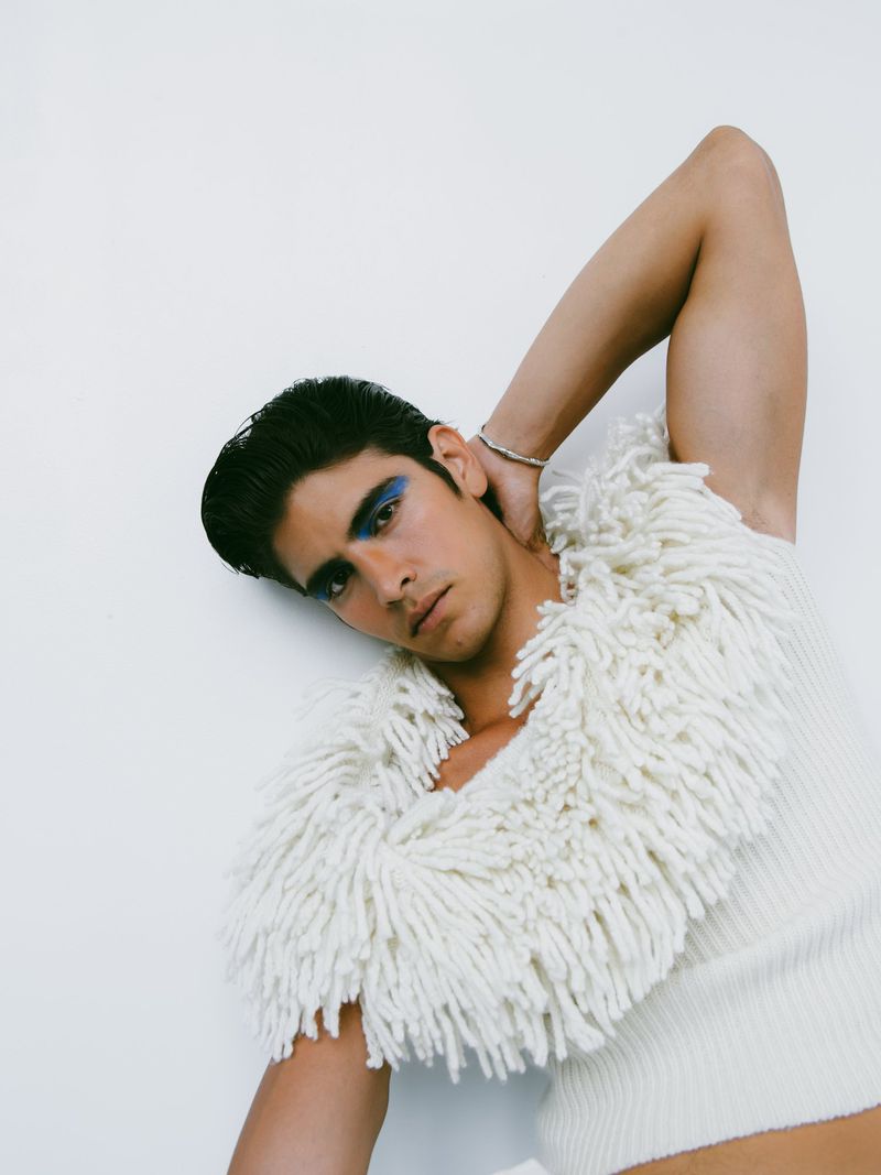 Man poses in a studio wearing a fuzzy off-white top with blue eyeshadow. He has dark hair and his right arm raised. 