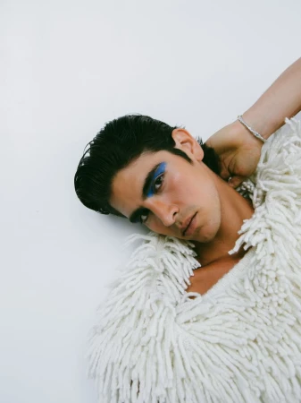 Portrait of adult male with dark hair and blue eyeshadow, wearing a shaggy white coat against a white background in a studio shot.