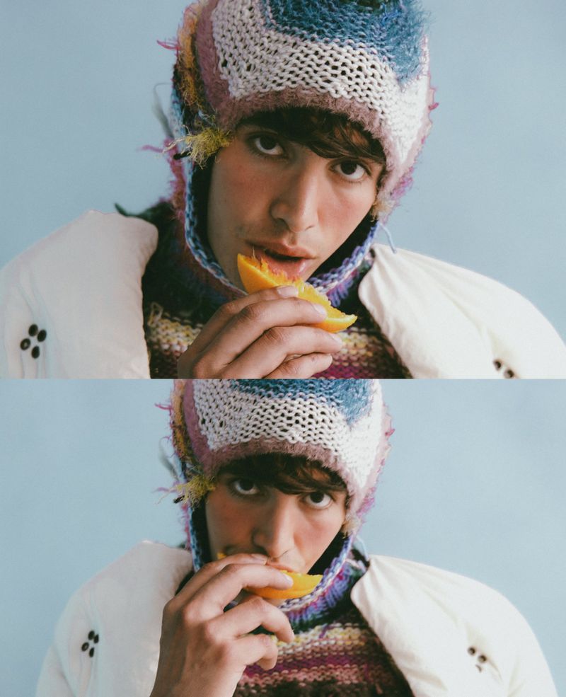 Young man in a colorful knit hat eats an orange slice against a light blue background, wearing a patterned sweater and white jacket.