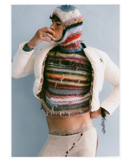 A person poses in a colorful knitted balaclava, top, and skirt with a white jacket while holding a tangerine in an indoor environment.