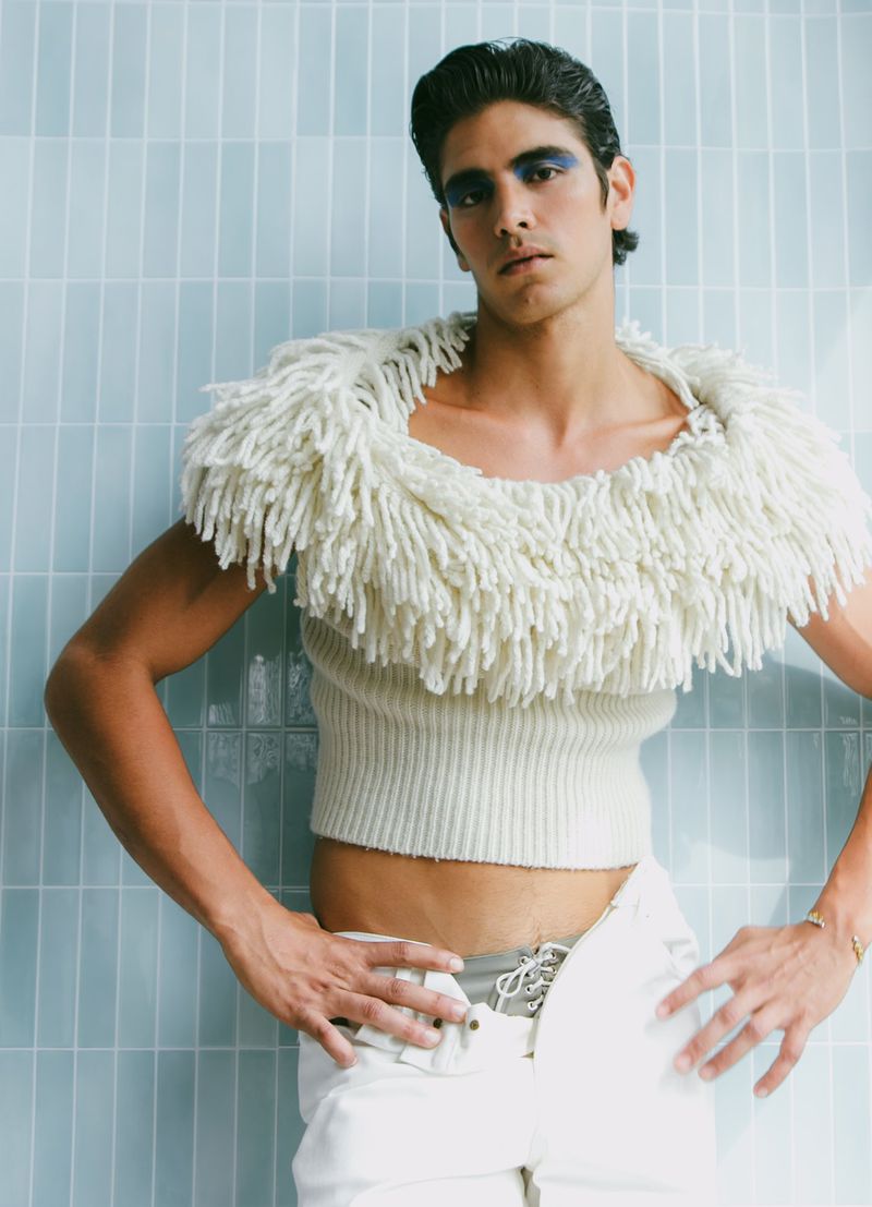 Fashion shot of a man posing in a cream-colored sweater with a fringe collar and white pants against a light blue tiled background.