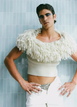 Fashion shot of a man posing in a cream-colored sweater with a fringe collar and white pants against a light blue tiled background.