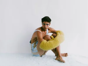 Model Sergio Perdomo seated on a white textured floor wearing a blue striped outfit and holding a fuzzy yellow prop for GQ Portugal