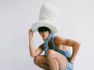 Male model wearing a large white top hat and blue striped knit bodysuit against a white backdrop.