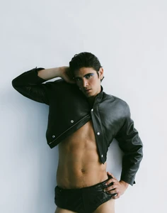 Model wearing a cropped black leather jacket and briefs against a plain white studio background