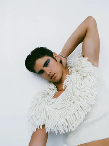 Male model wearing a white textured fringed top with blue eye makeup posing against a plain white background