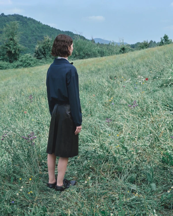 Model Meadow Walker in a dark navy jacket and dark skirt standing in a lush green meadow for Jil Sander