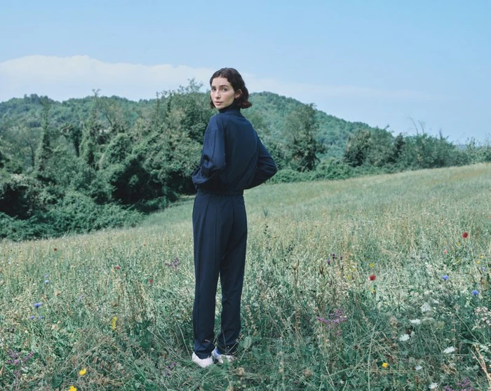 Model Meadow Walker in a dark navy blue matching track set standing in a lush grassy meadow for Jil Sander and Puma AW26.