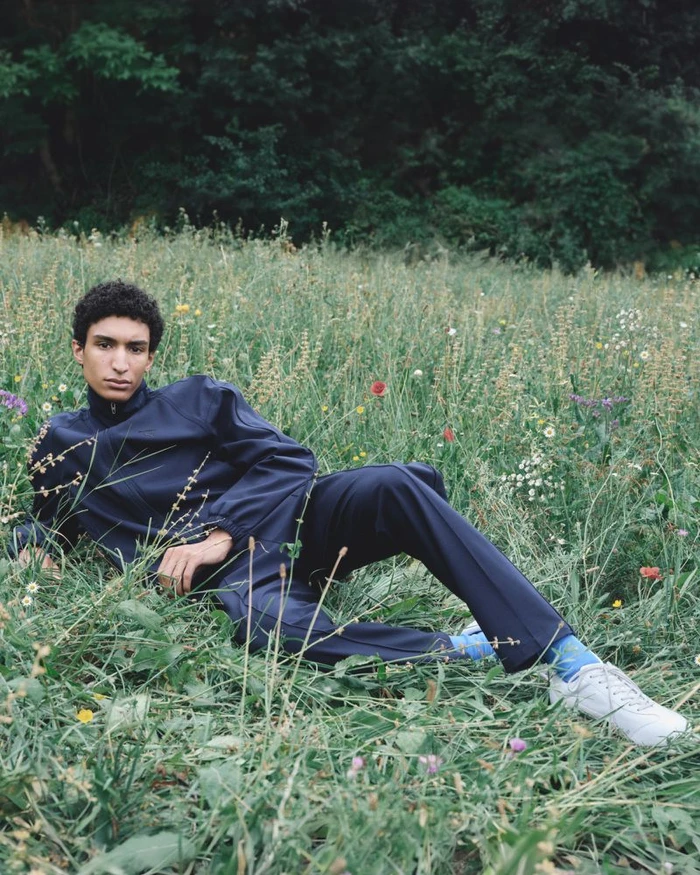 Man wearing a navy blue tracksuit reclining in a grassy wildflower field outdoors.