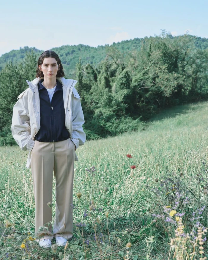 Model standing in a grassy meadow wearing an off-white jacket, dark zip-up top, and beige trousers