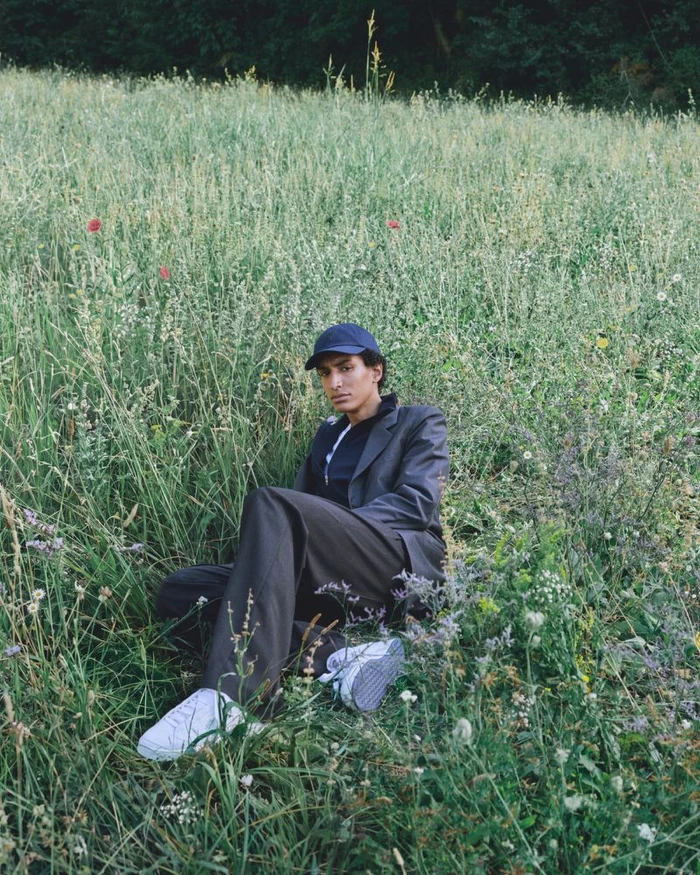 Model wearing a dark blazer, trousers, and baseball cap seated in a lush wildflower meadow.