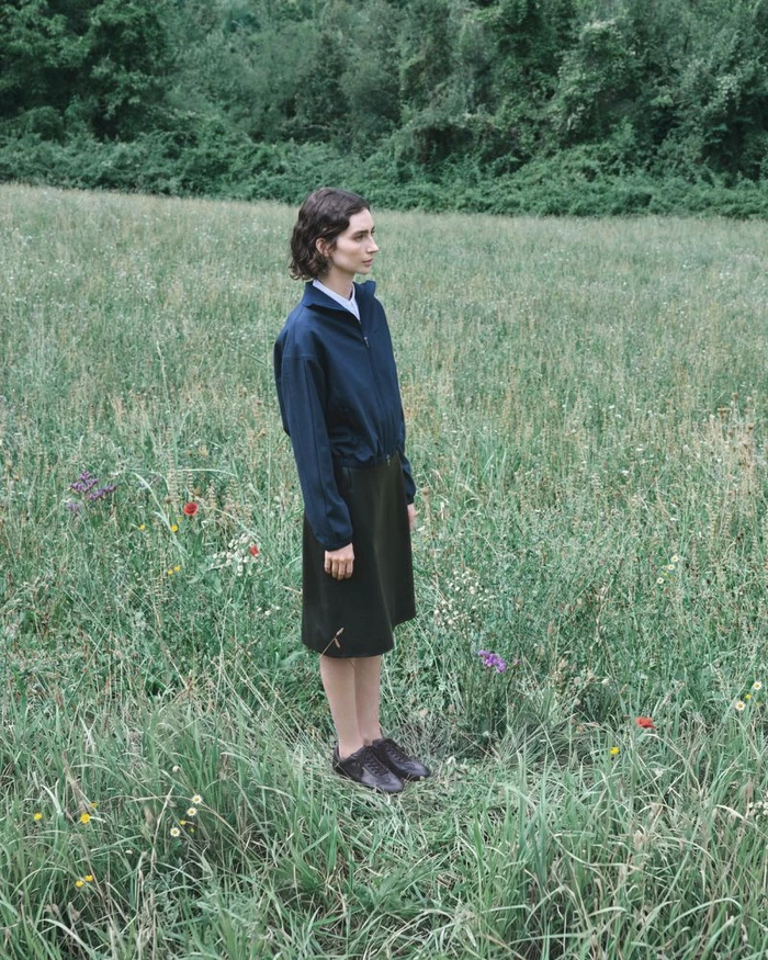 Meadow Walker standing in a lush green meadow wearing a dark zip-up jacket and midi skirt for Jil Sander.